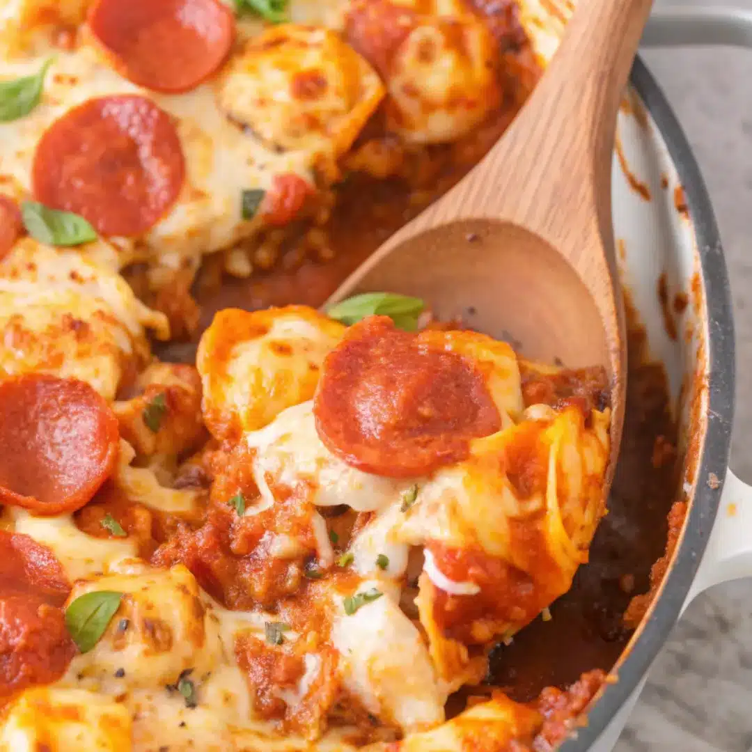 Lifting a Serving of Cheesy Pasta Bake Wooden spoon lifting cheesy tortellini and pepperoni from skillet